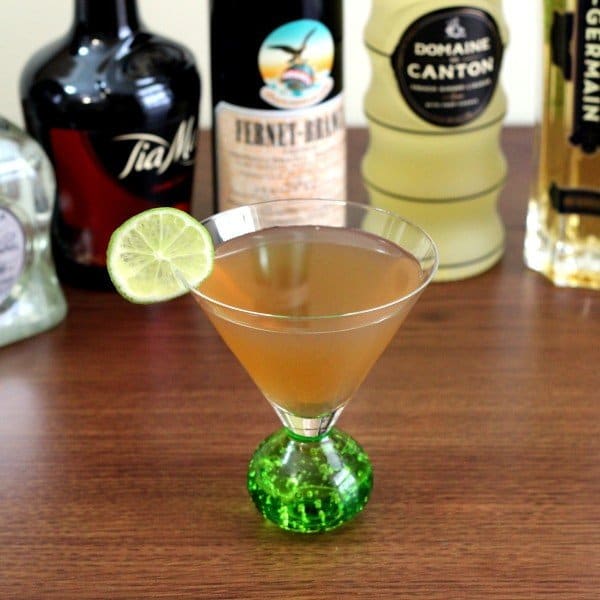 March Rain Cocktail with lime wheel in front of bottles of liquor