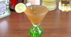 March Rain Cocktail with lime wheel in front of bottles of liquor