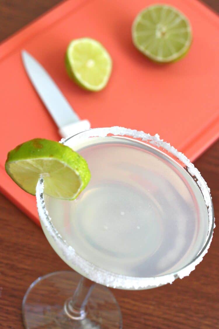 Overhead shot of margarita drink in glass beside cutting board with limes